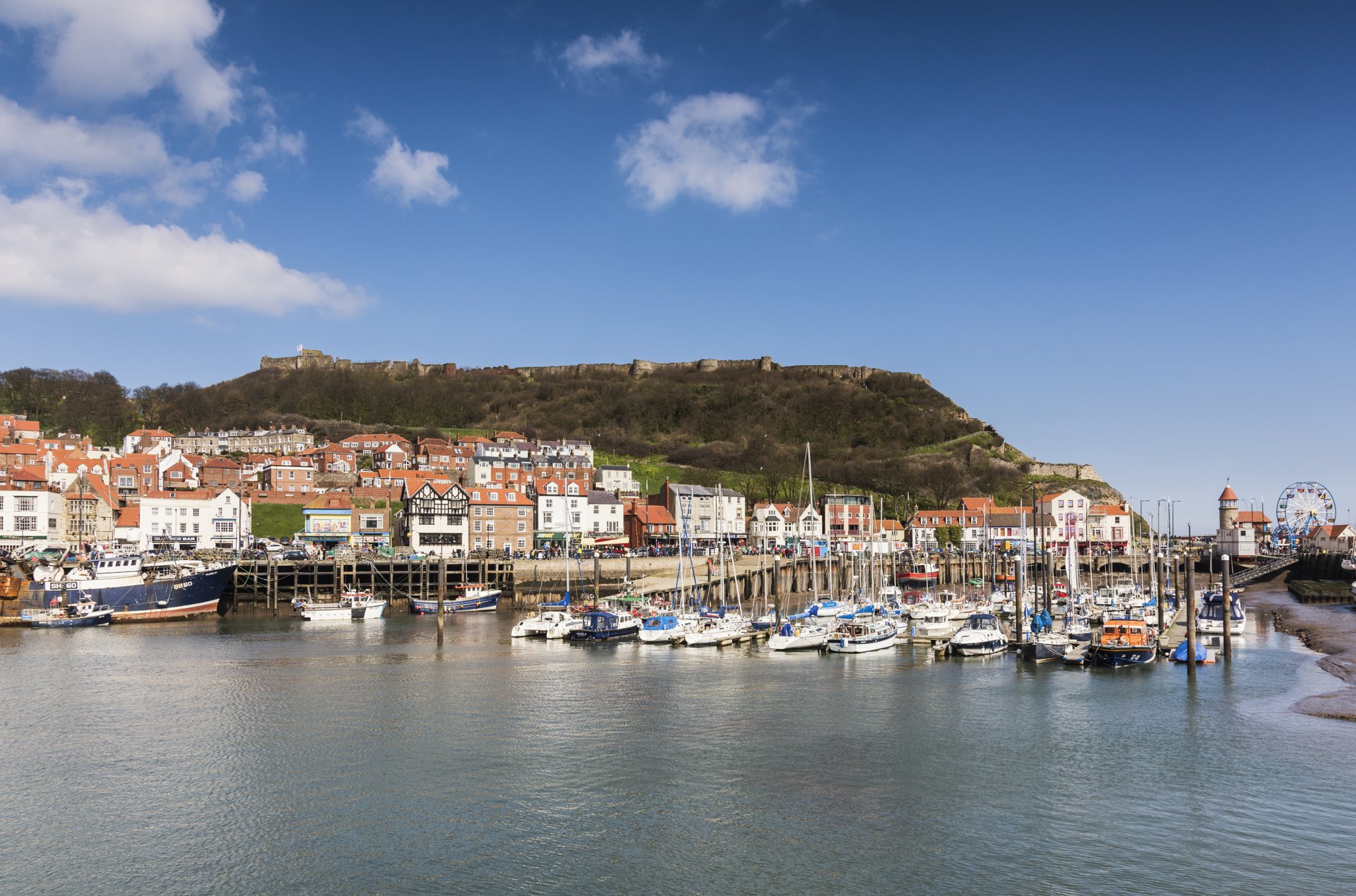 The harbour at Scarborough, a row of boats are moors on the dock, and in the background the outline of Scarborough Castle is visible against a clear blue slky